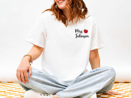 Person wearing a white t-shirt with 'Mrs. Johnson' and an apple design, sitting on a white couch.